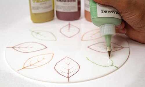 Person painting leaf design on ceramic plate with paint bottles in background.